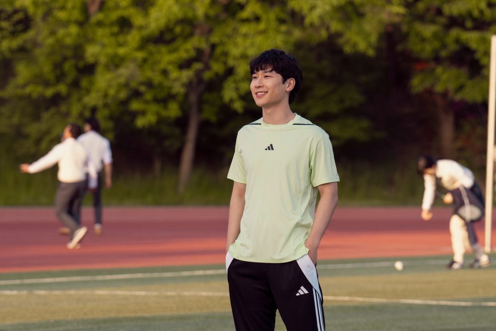 A young man stands on a sports field, smiling and looking off into the distance. He is wearing a light green athletic shirt and black Adidas track pants with white stripes, with his hands in his pockets. The background features a red running track and lush green trees. Other individuals are visible in the distance, engaged in activities like walking and practicing sports, adding to the casual and outdoor atmosphere.