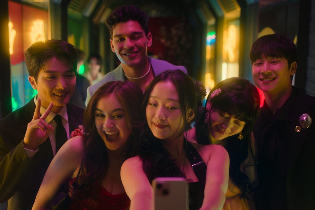 A group of six friends gather closely together, smiling and posing for a selfie in a lively and colorful setting. The central figure, holding the phone, wears a black dress with floral accessories, while others around her are dressed in formal attire, including a red dress, suits, and vibrant dresses with floral accents. One person flashes a peace sign, adding a playful touch. The background is illuminated with bright, neon-colored lights, creating a festive and energetic atmosphere. The group radiates happiness and camaraderie, capturing a joyful moment.