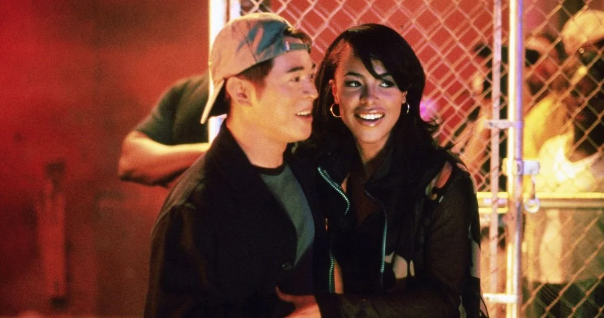 Aaliyah as Trish O’Day and Jet Li as Han Sing stand together in front of a chain-link fence, illuminated by warm, colorful lighting. The man, wearing a backward baseball cap, a dark jacket, and a gray shirt, smiles while looking off to the side. The woman, with long dark hair, hoop earrings, and a stylish black jacket with colorful accents, smiles brightly as they stand close together. The background features a lively, blurred crowd, adding to the energetic and urban atmosphere of the scene.
