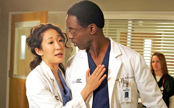 A scene set in a hospital features Dr. Cristina Yang (played by Sandra Oh) and Dr. Preston Burke (played by Isaiah Washington), both wearing white lab coats with ID badges. Cristina, with wavy dark hair, appears to be mid-conversation, placing her hand on Burke's chest with an expression of concern or urgency. Burke, wearing glasses with short-cropped hair, looks at her attentively with a serious expression. Both are dressed in scrubs under their lab coats. In the background, a nurse in black scrubs stands observing, with hospital blinds and a wooden door visible, indicating a clinical setting.