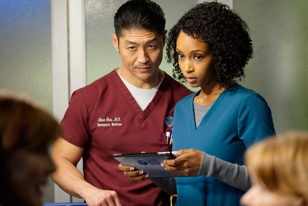 Two medical professionals, Dr. Ethan Choi (wearing maroon scrubs with "Emergency Medicine" embroidered on his uniform) and Dr. April Sexton (wearing teal scrubs and holding a tablet), are standing side-by-side in a hospital setting. Ethan Choi has a serious and focused expression, while April Sexton looks concerned as they both examine the information on the tablet. The background is softly blurred with other medical staff visible, creating an atmosphere of urgency and collaboration typical of a hospital environment.