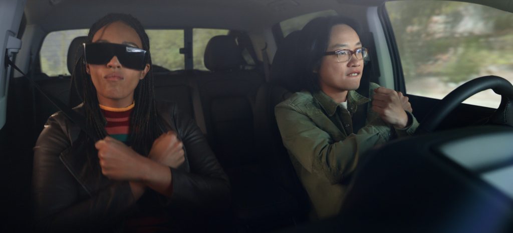 A scene from Space Force shows Jimmy O. Yang, portraying Dr. Chan Kaifang, driving a car with a determined expression, gripping the steering wheel tightly. He is wearing glasses and a green jacket with a slightly worn texture. Beside him in the passenger seat, Tawny newsome as Captain Angela with long braids, wearing a leather jacket and large futuristic visor-style sunglasses, crosses her arms in an assertive gesture. She wears a colorful striped sweater beneath her jacket. Both are wearing seat belts. The background outside the car window is blurred, showing trees and a road, indicating they are in motion. The scene has a playful and intense mood, highlighting the characters' expressions and body language.