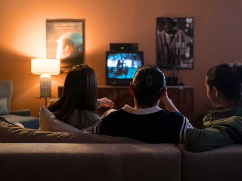 Back of the head of three friends sitting on a couch watching TV in dim lighting for Autism Acceptance Month.