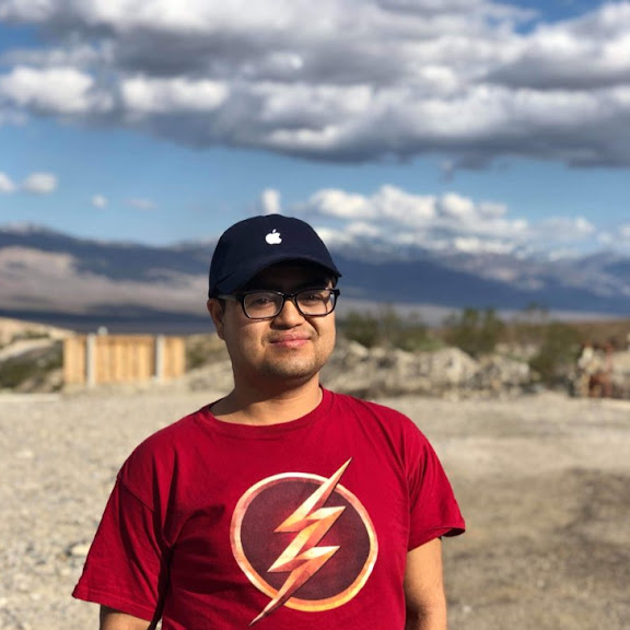 Dennis stands outdoors on a bright, sunny day with a mountainous desert landscape in the background. They are wearing a red Flash superhero T-shirt, a navy Apple baseball cap, and black-framed glasses. The person is smiling slightly, with a clear blue sky and scattered clouds behind them.