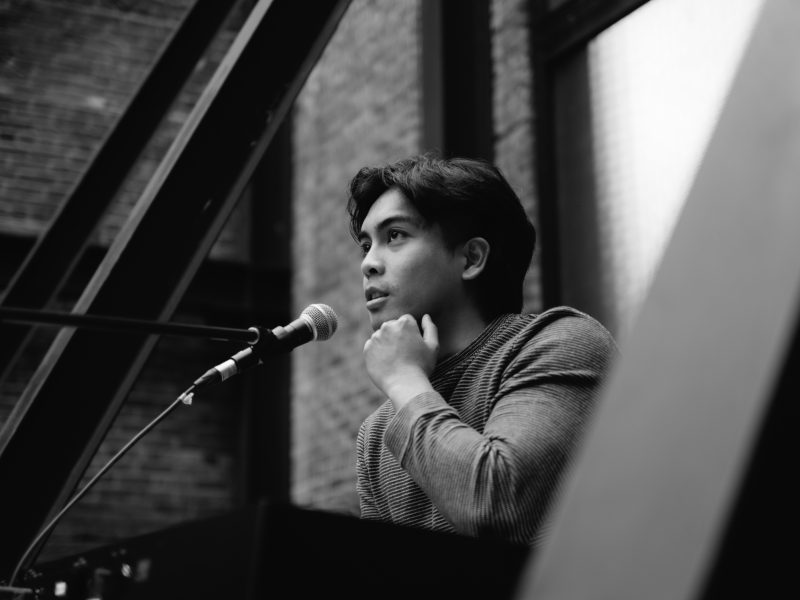 Black and white photo of music artist Mar Emanuel looking to the left with his hand under his chin and microphone in front of his mouth.