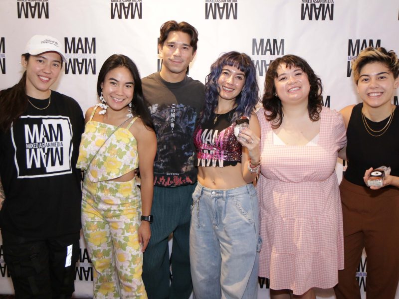 A group of seven individuals, mostly women, gather closely and smile for a photo at an event. The group features sparkly dresses, floral prints, and stylish casualwear. The background shows a projected screen and dim lighting, suggesting an evening event or showcase. Two people kneel in front while others stand behind them with arms around each other, creating an intimate and celebratory energy.