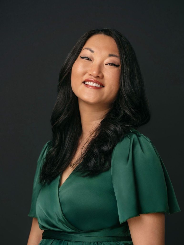 A portrait of Grace Yung Foster smiling warmly against a dark background. She has long black wavy hair and wears a deep green dress with short flutter sleeves. The lighting highlights her face and confident expression, creating a professional and welcoming tone.