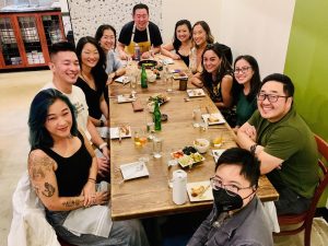 A group of twelve people gather around a long wooden dining table, smiling for the camera at a restaurant. Plates of food and bottles of drinks are arranged across the table, and the atmosphere feels lively and joyful.