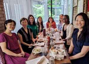 A group of nine people sit together around a long table in a restaurant, smiling at the camera. Plates of food, glasses of water, and wine are visible on the table. Natural light streams in from large windows behind them, creating a warm and welcoming atmosphere.