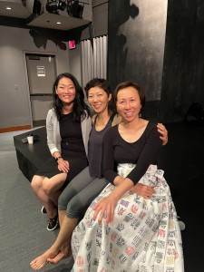 Three women sit together on a small theater stage, smiling and posing for a photo. Behind them, the dark walls of the stage are visible. One woman wears a skirt with colorful calligraphy-style designs, another wears a black top and gray pants, and the third wears a blazer and sneakers.