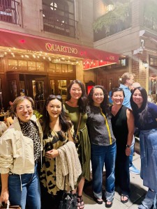Seven people pose outside a restaurant with a red awning that reads “Quartino.” They stand close together, smiling warmly at the camera in casual clothing. The restaurant’s string lights glow behind them, adding to the celebratory evening mood.