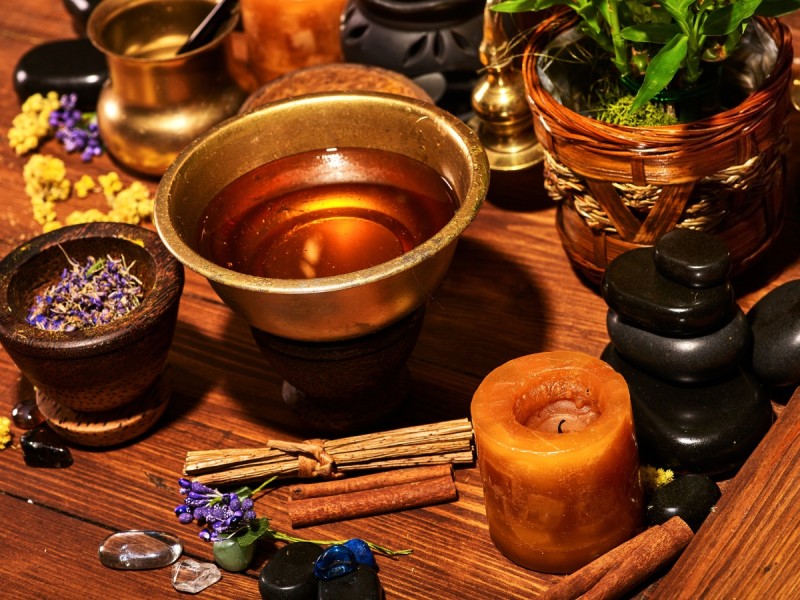 A warm, spa-like still life on a wooden table featuring a brass bowl filled with amber-colored herbal oil, surrounded by smooth black stones, dried lavender and yellow flowers, cinnamon sticks, a small candle, crystals, and woven baskets with green plants, evoking a calming Ayurvedic wellness ritual.
